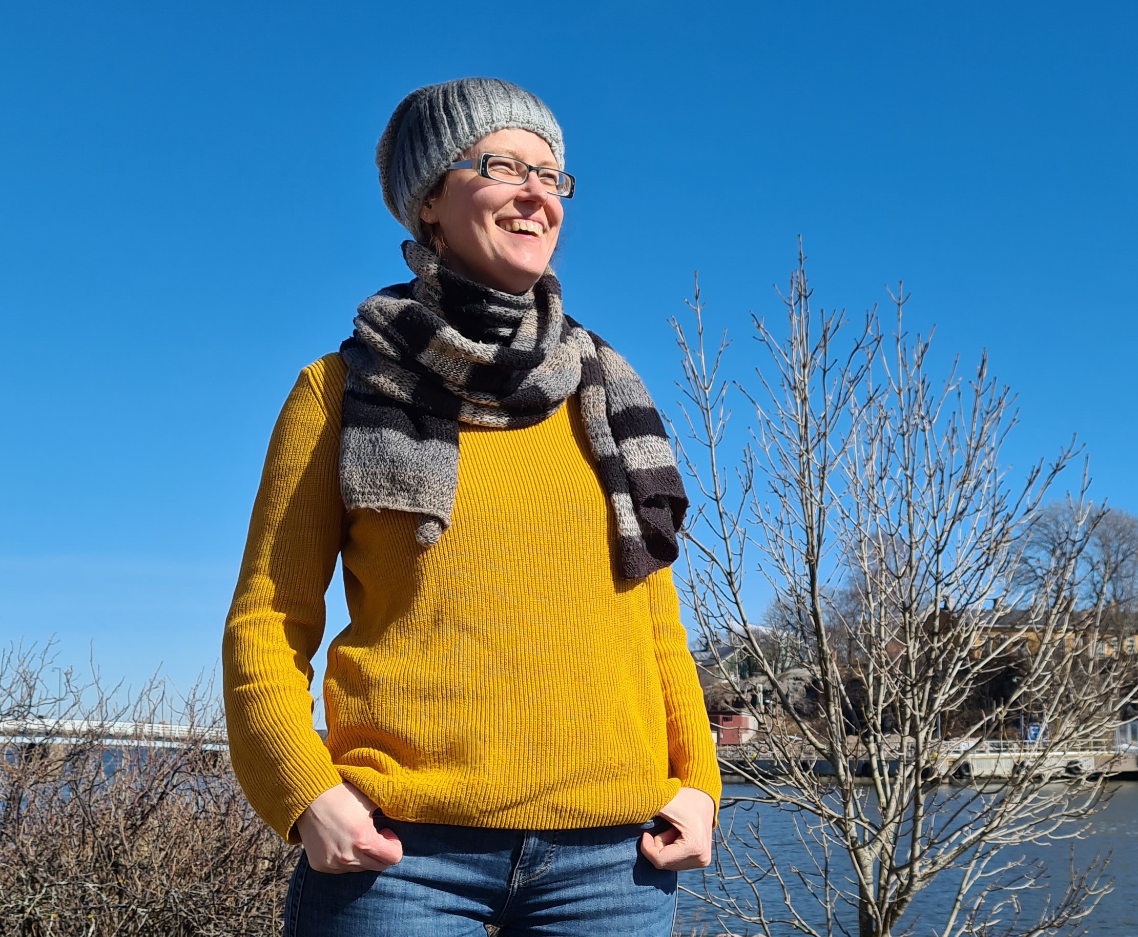 Das Foto zeigt eine junge Frau in Jeans und gelbem Pullover mit Schal und Mütze, die in lockerer Pose draußen vor einem natürlichen Hintergrund unter blauem Himmel steht. Sie trägt eine schwarze Brille und lacht. Im Hintergrund sind einige Sträucher und Wasser zu sehen.